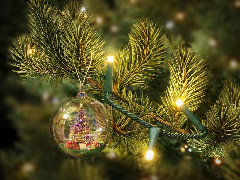 Close-up van donkergroene kerstboomtakken met warmwitte bullet-head LED-verlichting en een glazen kerstbal met daarin een miniatuur kerstboom en rode geschenken.