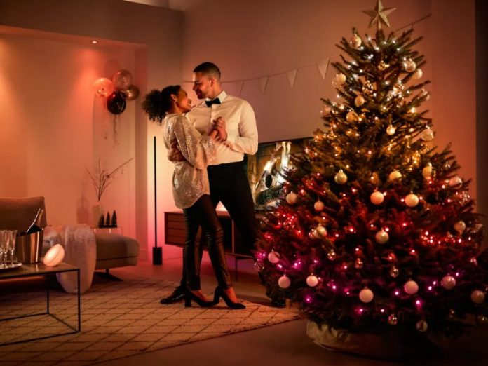 Een stel danst romantisch in een sfeervol oranje verlichte woonkamer, met een kerstboom versierd met warme witte en roze lampjes en gouden kerstballen.