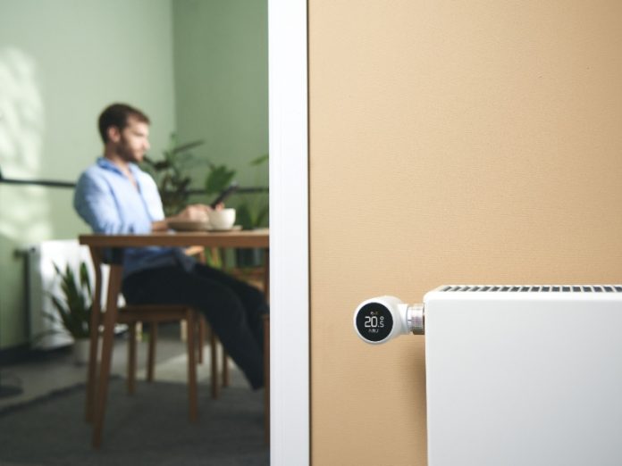 Witte Tado slimme thermostaatknop op een radiator in een lichtbeige kamer, toont 20.5 graden. In de zachtgroene achtergrond zit een onscherpe man in een lichtblauwe blouse aan een houten tafel.