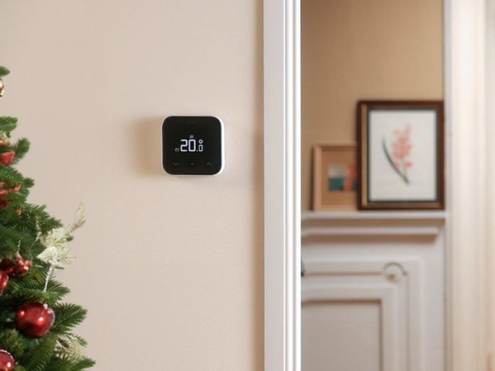 tado-11 Witte tado thermostaat met 20.0° op een lichtbeige muur, naast een groene kerstboom met rode ballen. Op de achtergrond opent een persoon in een beige jas een witte deur naar een hal met een rode jas en ingepakte kerstcadeaus.