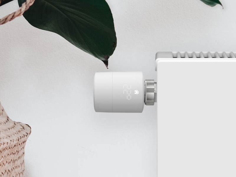Witte Tado slimme thermostaatknop toont 22.0 graden op een witte radiator, met donkergroene plantenbladeren en een lichtbeige rieten mand in een modern interieur.