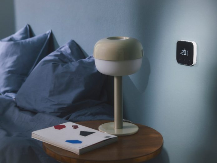 Witte Tado slimme thermostaat met display '20.0°' op een lichtblauwe muur, naast een beige lamp en donkerblauw beddengoed.