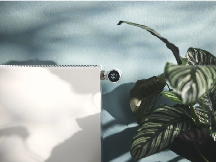 Witte radiator met een digitale tado slimme radiatorknop die 20,5 graden aangeeft, geplaatst tegen een lichtblauwe muur met zachte schaduwen van bladeren naast een gestreepte groene kamerplant.