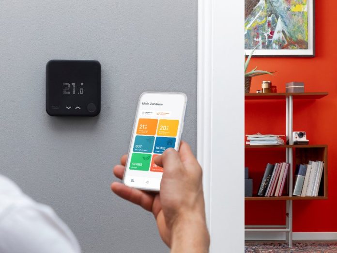 Zwarte Tado thermostaat op een lichtgrijze muur, een hand toont de Tado app op een smartphone, met op de achtergrond een oranjerode kamer met een witte radiator en kerstdecoratie.