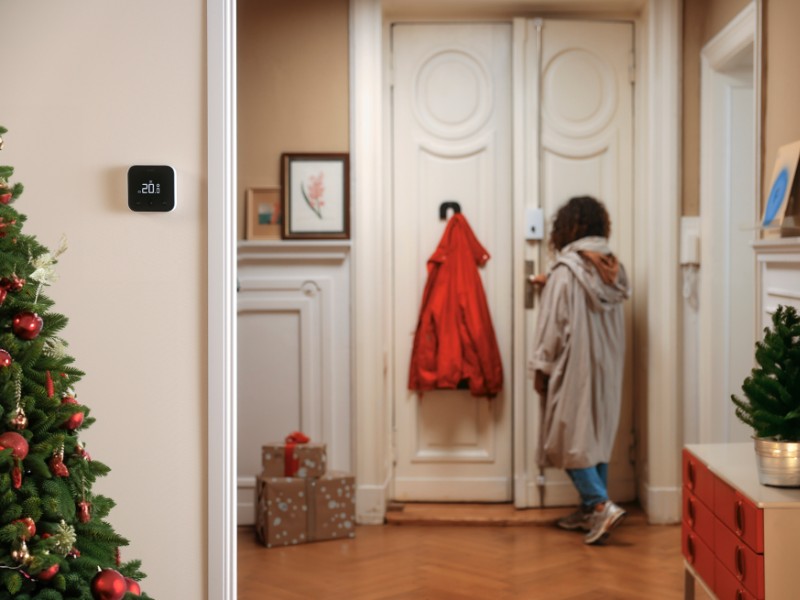 Een zwarte tado thermostaat, gemonteerd op een licht beige muur, toont 20.0 graden Celsius. Links staat een groene kerstboom met rode kerstballen, wat een warme en feestelijke sfeer creëert in de hal.