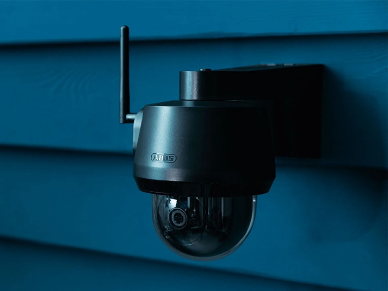 Doffe, donkergrijze ABUS domecamera met antenne, gemonteerd op een donkerblauwe houten gevel.