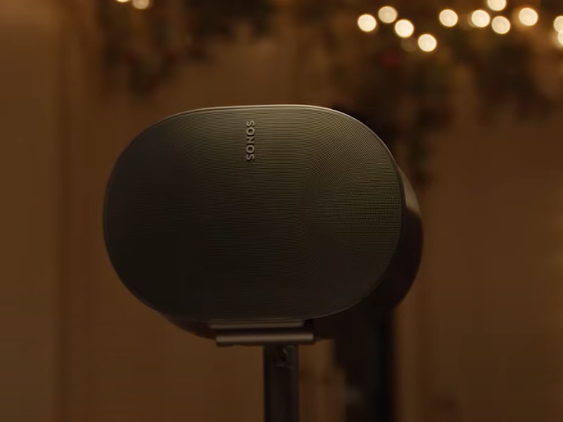 Donkergrijze Sonos luidspreker op een standaard, met op de achtergrond wazige, warme kerstverlichting, relevant voor Huis beveiligen feestdagen.