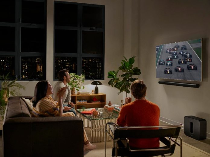 Drie opgewonden kijkers bekijken een felverlichte autorace op tv in een donkere woonkamer, ondersteund door een zwarte Sonos thuisbioscoop-opstelling met soundbar, Sub en achterspeakers.