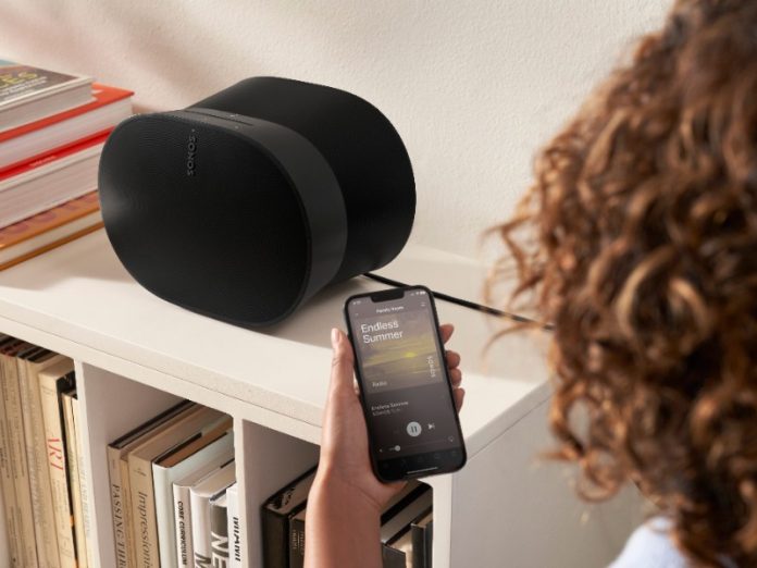 Hand met zwarte smartphone toont de Sonos app, gericht op twee matzwarte Sonos-luidsprekers op een witte boekenplank.