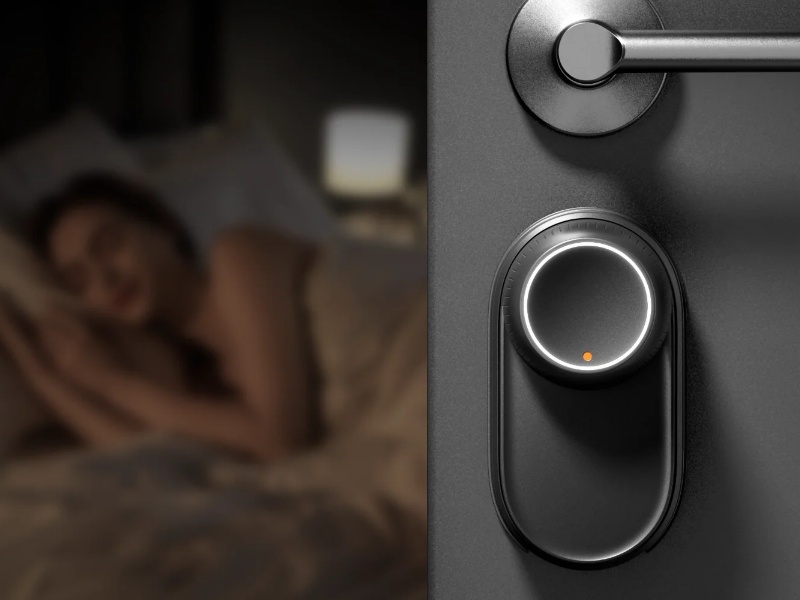 Dichrome afbeelding van de SwitchBot Lock Ultra in nachtmodus op een donkere deur naast een zilveren klink, met een oranje indicatielampje; links ligt wazig een slapende persoon in bed. De sluishaak genereert minder dan 20dB geluid.