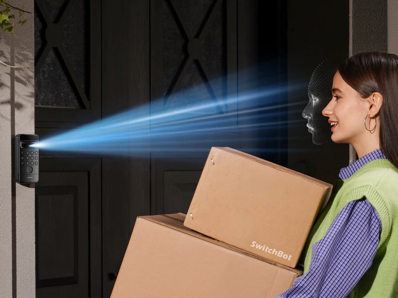 Zwarte SwitchBot Lock Ultra op een bakstenen muur, zendt een blauwe lichtstraal uit voor 3D-gezichtsherkenning van een jonge vrouw die kartonnen dozen vasthoudt.