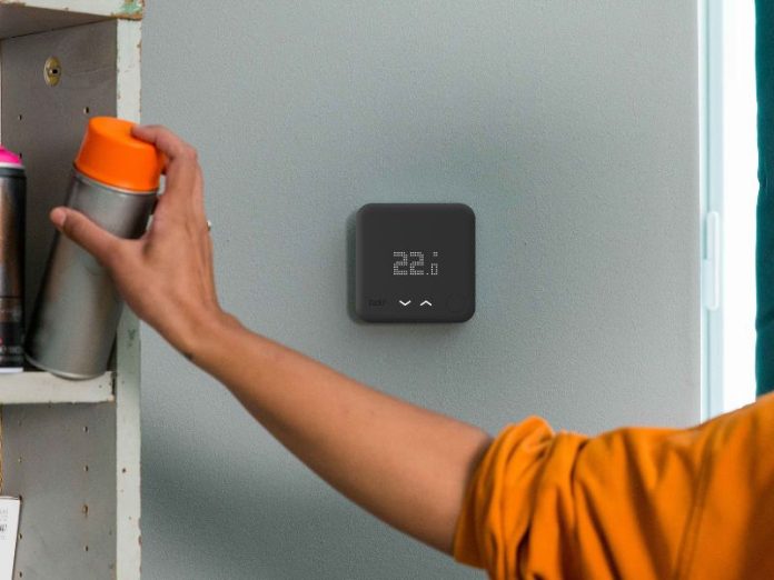 Een persoon in een oranje shirt reikt naar een spuitbus met een oranje dop op een houten plank naast een zwarte tado slimme thermostaat op een grijze muur, die een temperatuur van 22,1 graden aangeeft.