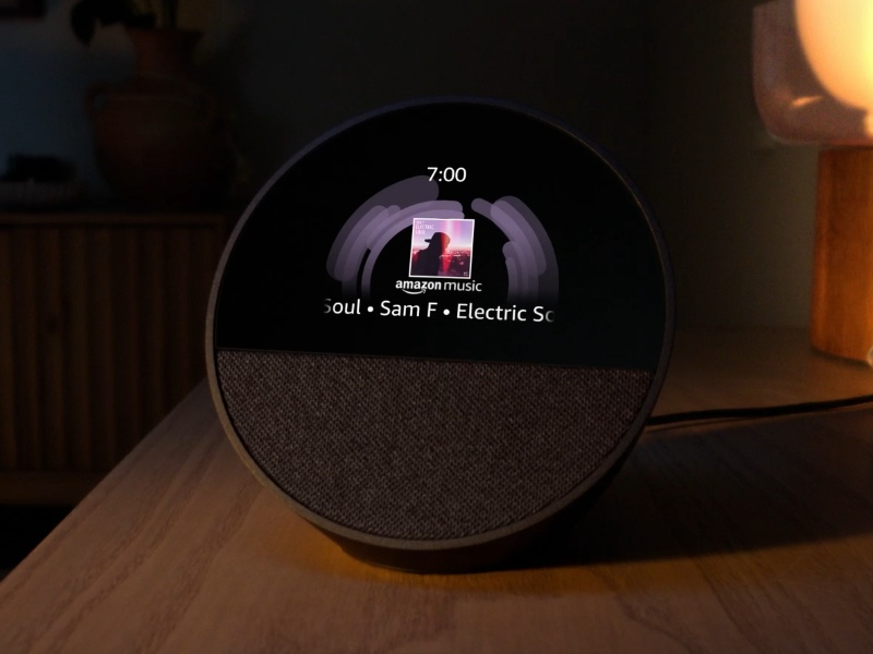 Donkergrijze Amazon Alexa smart display op een houten tafel in een schemerige kamer, toont paarse audioweergave en albumkunst van Amazon Music.