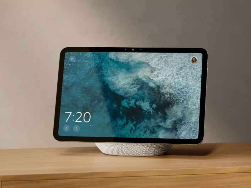 Zwart smart display van Amazon Alexa op een lichte houten kast. Het scherm toont turquoise golven en de tijd 7:20, en rust op een witte geweven luidsprekerbasis.
