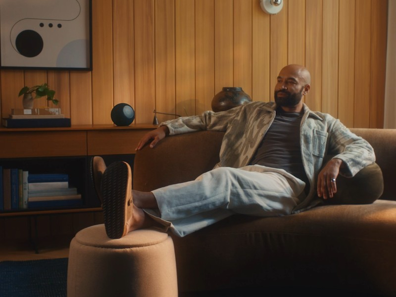 Man ontspant op een bruine fluwelen bank in een warm, met hout bekleed interieur, met een zwarte Amazon Alexa-speaker op het dressoir.