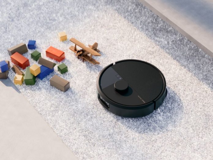 Zwarte robotstofzuiger (lijkend op een Roomba) op een lichtgrijze, hoogpolige mat, omringd door gekleurde houten speelgoedblokken en een houten vliegtuigje. Dit illustreert de uitdagingen na het iRobot faillissement Roomba.