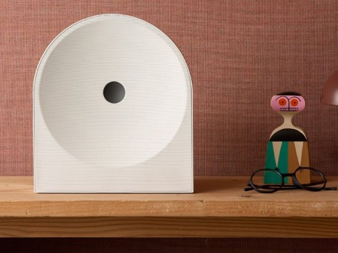 Gebroken witte, halfronde Samsung Audio 2026 luidspreker met donkergrijs accent op een houten plank, naast een kleurrijke houten pop en zwarte bril tegen een roodbruine muur.