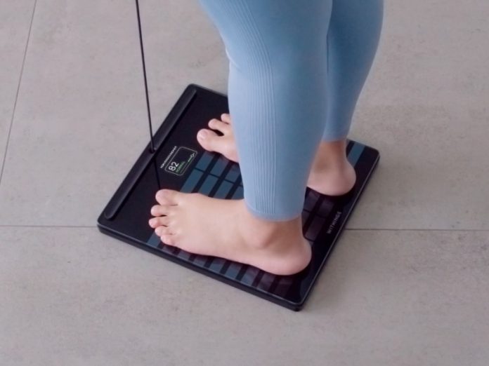 Voeten op een zwarte, glanzende Slimme weegschaal goede voornemens, met een groene meting van '82 NORMAL' zichtbaar op het display, terwijl de persoon een lichtblauwe legging draagt op een grijze tegelvloer.