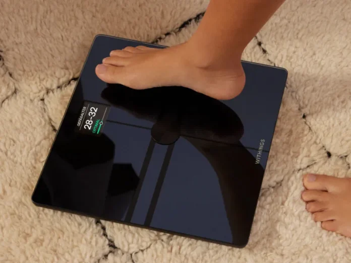 Detail van een donkere, glanzende Withings slimme weegschaal met een blote voet erop, geplaatst op een witte, pluizige mat. Het scherm toont een 'OPTIMAL' resultaat in groen, behorend tot de Beste slimme weegschalen 2026.