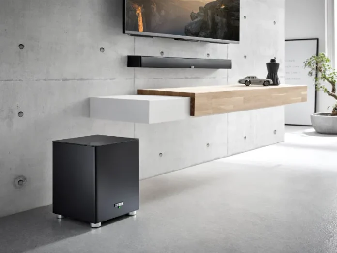 Image: Zwarte Canton subwoofer op de vloer in een moderne ruimte met TV, soundbar en zwevende plank tegen een grijze betonnen muur.