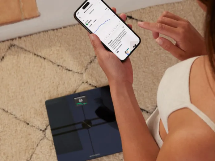 Close-up van een hand die een smartphone bedient, waarop gezondheidsdata te zien is, terwijl op de vloer een donkerblauwe slimme weegschaal ligt met de weergave '58'. Dit toont de connectiviteit van de Beste slimme weegschalen 2026.