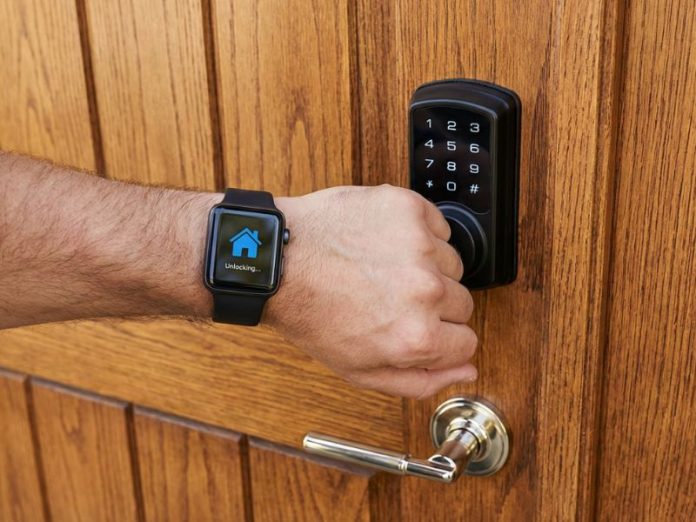 **Apple HomeKey uitleg:** Een arm met een Apple Watch wordt gebruikt om een slim slot op een houten deur te ontgrendelen, waarbij de smartwatch de status ‘Unlocking...’ toont.