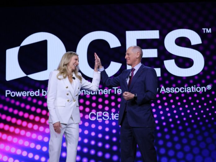Vrouw in wit pak met gouden knopen en man in marineblauw pak geven high-five op het CES 2026 Recap podium voor een scherm met het witte CES-logo en magenta verlichting.