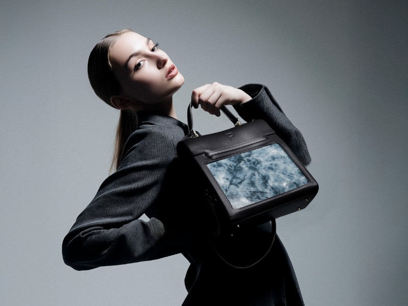 Jonge vrouw in donkergrijze blazer poseert met een zwarte, futuristische handtas met een vierkant, marmerachtig blauw-wit display, inspelend op CES 2026 verwachtingen.