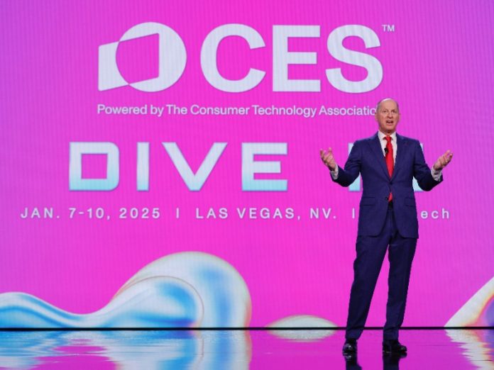 Man in donkerblauw pak en rode das op podium met reflecterende vloer, sprekend voor een fel magenta CES scherm. Relevant voor CES 2026 verwachtingen.