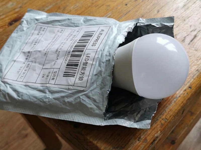 Grote witte LED-lamp steekt uit een gescheurde, grijze AliExpress verzendzak met Chinese labels, liggend op een versleten houten tafel.