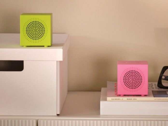 Twee felgekleurde IKEA smart home kubusluidsprekers (limoengroen en felroze) op een witte plank, getoond op CES 2026, naast een grote zwarte bril.