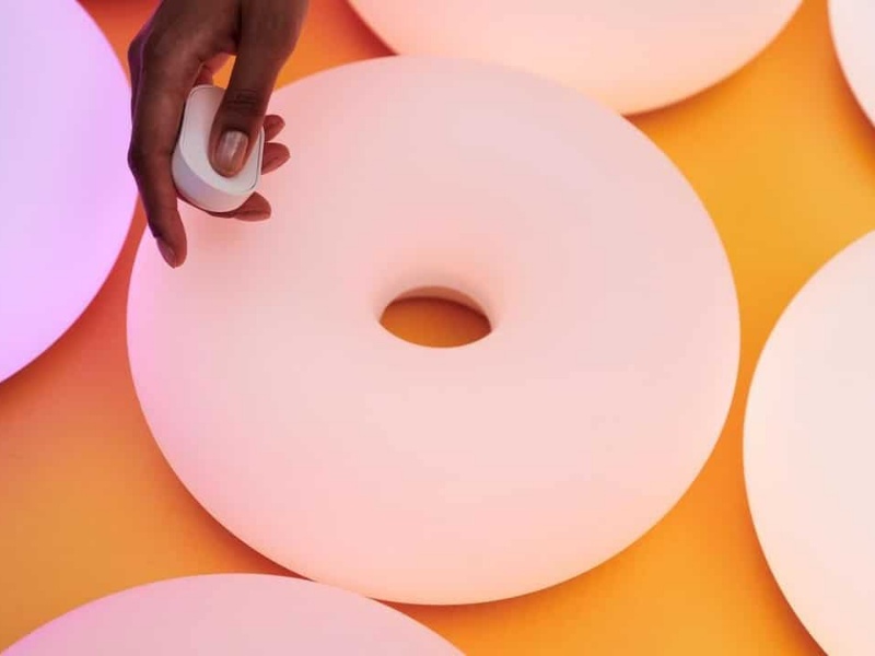 Donkere hand met witte afstandsbediening boven lichtgevende, torusvormige lampen in roze en paarse tinten op een oranje ondergrond, IKEA smart home CES 2026.