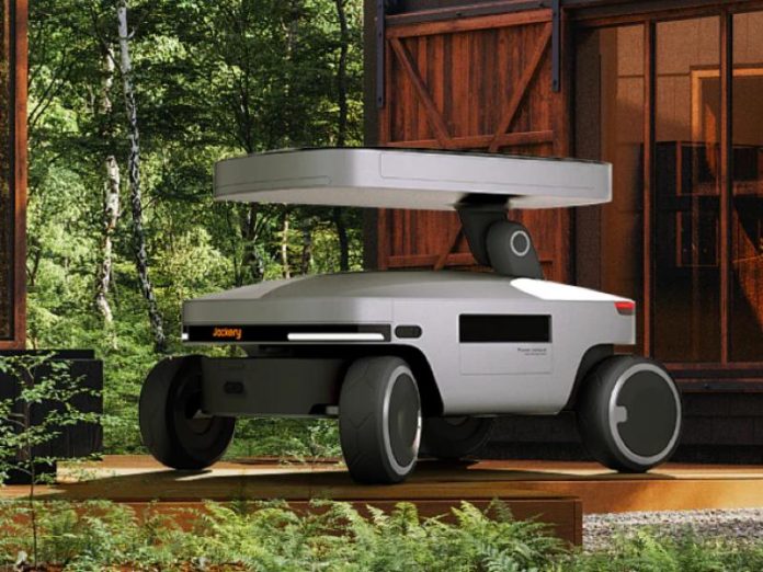 Grijze Jackery Solar Mars Bot op vier zwarte wielen met wit zonnedak, geparkeerd op een houten terras voor een donkerbruin hutje in een groene bosrijke omgeving.