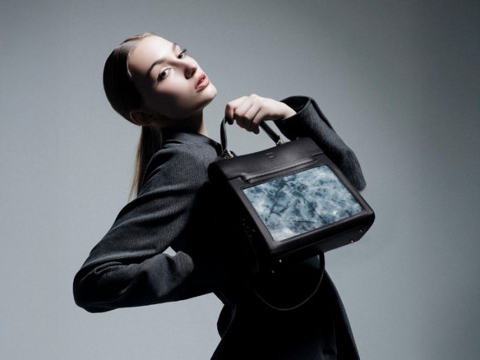 Model in een donkergrijze jas poseert met de zwarte Peuty Infinity OLED Handtas, waarvan het scherm een dynamisch lichtblauw marmerpatroon toont.