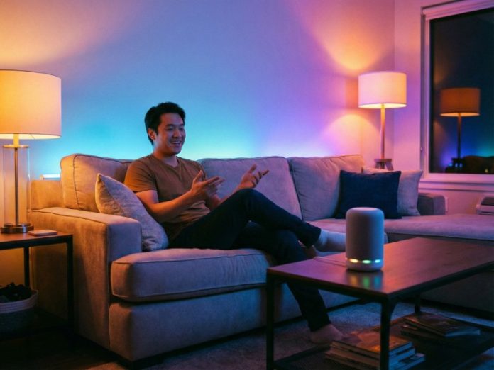 Lachende man op een sofa die gebaren maakt naar een witte slimme speaker op een salontafel, terwijl de woonkamer is verlicht met levendig blauw en paars smart lighting. Dit illustreert SmartThings spraakbesturing koppelen.