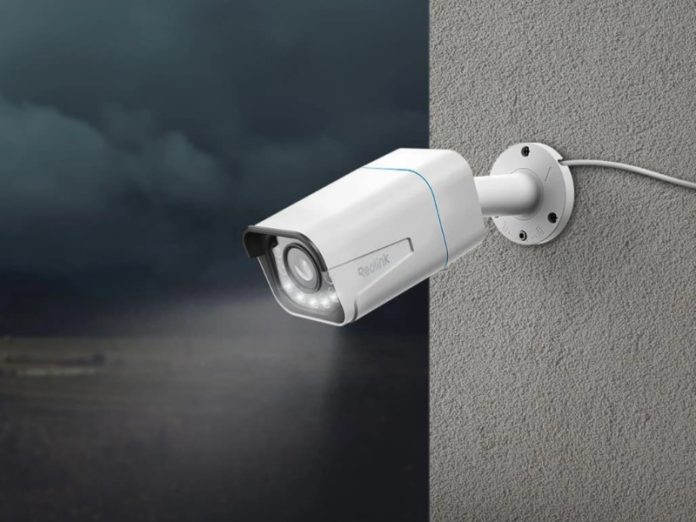 Witte Reolink beveiligingscamera (Beveiligingscamera zonder abonnement) gemonteerd op een grijze muur, waarbij het licht schijnt op een donker, wazig landschap met een afgelegen boerderij.