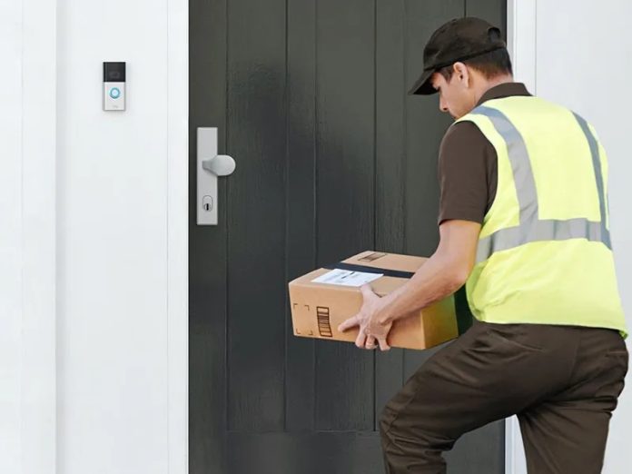 Een pakketbezorger, gekleed in een neongeel hesje, staat met een pakket voor een donkergrijze deur naast een zilverkleurige slimme video Deurbel.