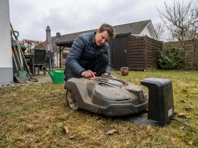 Een man in een donkerblauwe jas hurkt om een vuile donkergrijze Husqvarna robotmaaier te inspecteren in een tuin met dor gras, passend bij de vraag 'Robotmaaier start niet na de winter?'