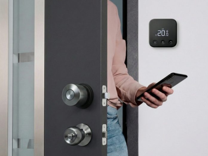 Een donkergrijze deur met geborstelde metalen sloten, waarbij een persoon met een zwarte smartphone binnenkomt. Op de witte muur ernaast hangt een zwarte slimme thermostaat die 20,0° aangeeft, illustrerend slimme verwarming en sloten.