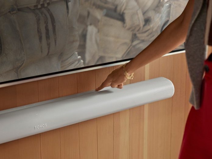 Witte Sonos soundbar gemonteerd onder een scherm op een houten wand, waarbij een hand de touchbediening aanwijst.
