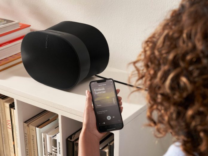 Hand bedient via de Sonos app op een smartphone een zwarte Sonos speaker, die op een witte plank naast boeken staat. De app speelt de track 