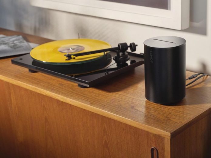 Zwarte platenspeler met felgeel vinyl op een houten dressoir, aangesloten op een cilindervormige zwarte Sonos-speaker.