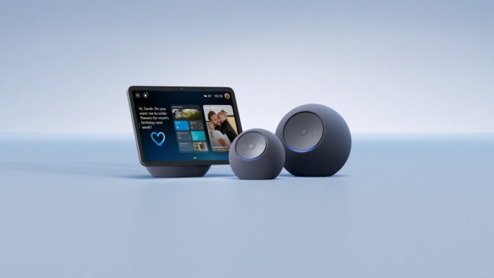 Een Amazon Alexa+ smart display en twee antracietgrijze bolvormige luidsprekers met blauwe lichtringen tegen een lichtblauwe achtergrond.