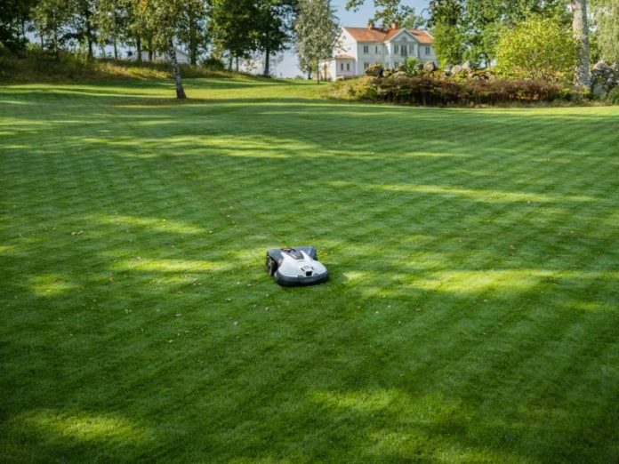 Een grijze robotmaaier op een uitgestrekt, strak groen gazon met een wit landhuis op de achtergrond, de beste robotmaaier voor een grote tuin.