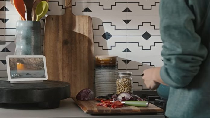 Handsfree koken met een wit smart display op een zwart plateau in een keuken, terwijl een persoon in een groenblauw shirt rode ui en paprika snijdt voor een wand met geometrisch patroon.