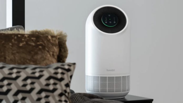 Een witte Hombli luchtreiniger met een groen verlicht display staat op een zwart nachtkastje als slimme hooikoorts hack. Op de voorgrond liggen bruine en zwart-wit gedessineerde kussens op een grijs bed.
