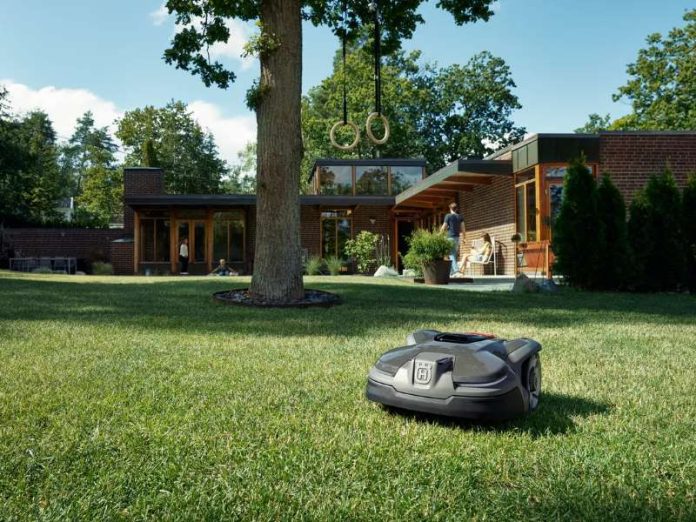 Een grijze Husqvarna robotmaaier op een groot, zonnig groen gazon voor een modern bakstenen huis.