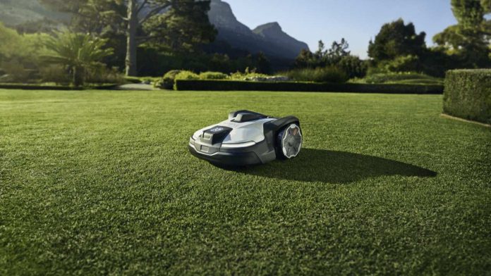 Een witte en grijze Husqvarna Automower robotmaaier aan het werk op een uitgestrekt groen gazon met bergen op de achtergrond, ter illustratie van de Husqvarna vs Segway Navimow vergelijking.