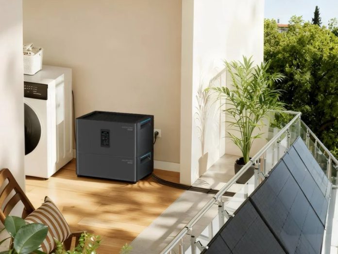 Een donkergrijze Marstek thuisbatterij op een zonnig balkon, aangesloten op zonnepanelen aan de reling en geplaatst naast een witte wasmachine.