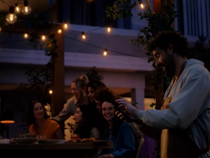 Een vrolijke groep vrienden geniet 's avonds buiten van een maaltijd, verlicht door warm amberkleurig licht van Philips Hue buitenverlichting. Een lachende man met baard draagt een bruin schort, terwijl meerdere vrouwen glimlachend rond een tafel zitten.